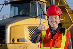 A royalty free image from the construction industry of a positive white collar construction engineer giving the thumbs up signal to show success. The worker is smiling and friendly standing in front of construction machinery which is a hauling truck. She is wearing safety glasses and a safety vest.